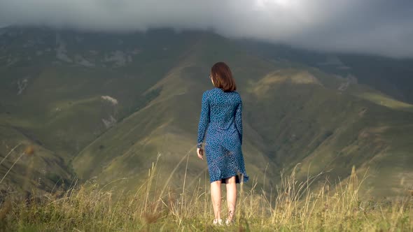 A Young Woman Stands in a Blue Dress and Raises Her Hands Up Looking at the Mountains.