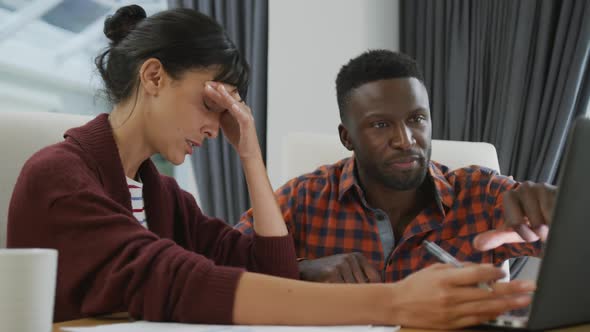 Diverse couple sitting at table talking and working with laptop