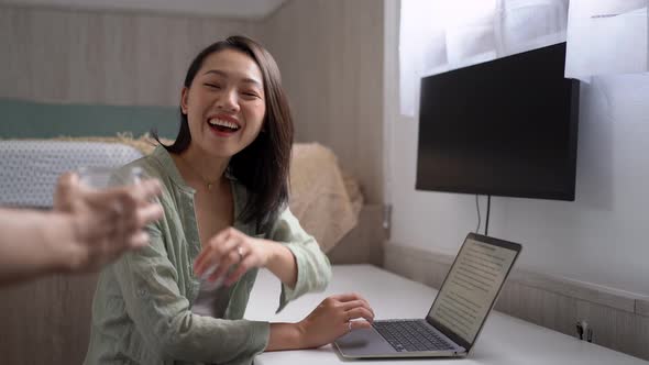 Crop partner passing cappuccino to smiling Asian blogger at home