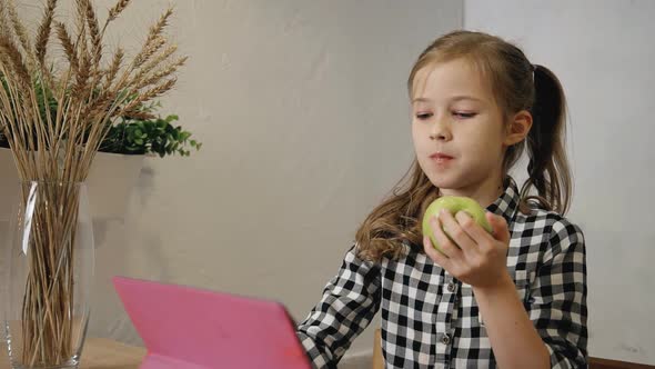 Little Girl Eating Apple and Using Tablet Computer in the Kitchen.