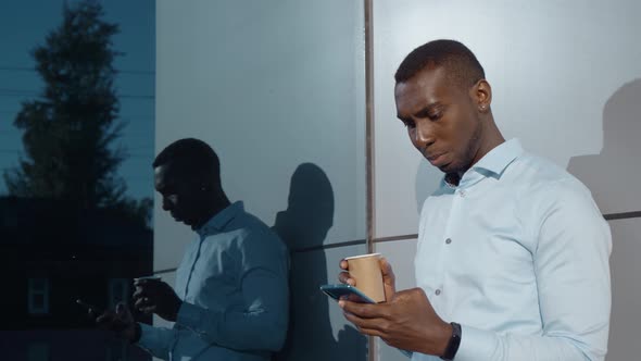 Afro American Man Is Drinking Coffee in Street at Morning, Viewing News Tape in Smartphone