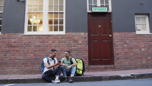 Two mixed race male friends sitting, talking and using smartphone in the street