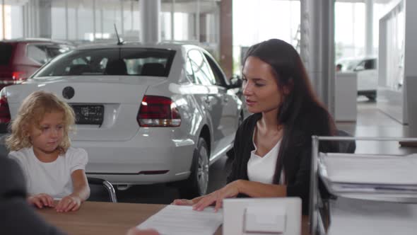 Woman Sitting with Daughter in Car Dealership and Signing Contract