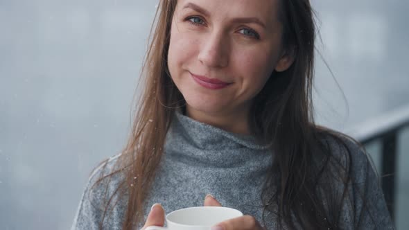 Caucasian Woman Stays on Balcony During Snowfall with Cup of Hot Coffee or Tea