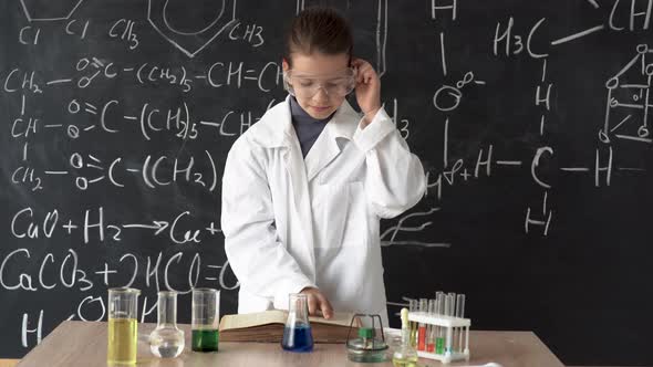 A Little Girl Scientist in a White Coat and Goggles Examines a Test Tube with a Chemical Reagent alt