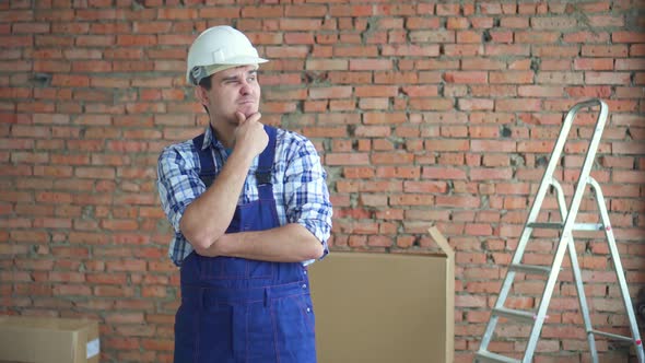 Puzzled Man in a Working Uniform and White Helmet Against a Brick Wall