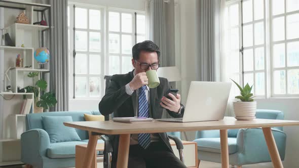 Asian Businessman Drinking Coffee And Using Mobile Phone While Working with Computer at Home