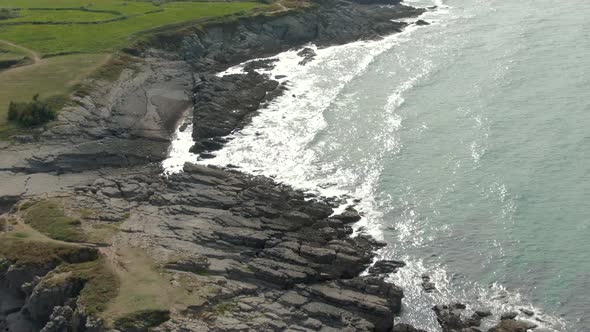 A drone rises slowly, over  the sea as the waves crash relentlessly against  jagged grey rocky shore