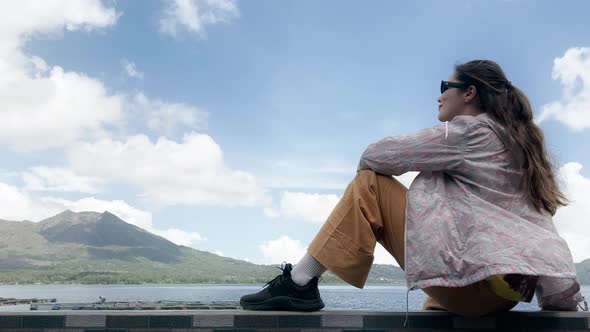 A Girl with Long Dark Hair Wearing Sunglasses a Yellow Pants is Sitting on Shore of Lake with