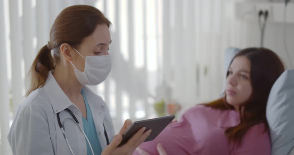 Female Doctor in Safety Mask Discussing with Pregnant Woman Over Digital Tablet in Hospital