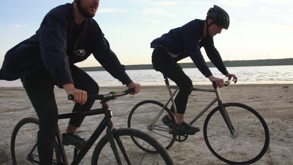 Two Young Men Riding Bicycles on the Beach on the Background of an Orange Sunsetting Sky