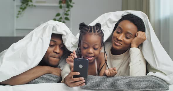Afro American Funny Family Lying on Sofa Bed at Home Under White Blanket Using Mobile Phone for