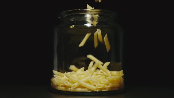 SLOW MOTION: Uncooked Penne Rigate Fall Into Empty Glass Jar On Black Background - Front View