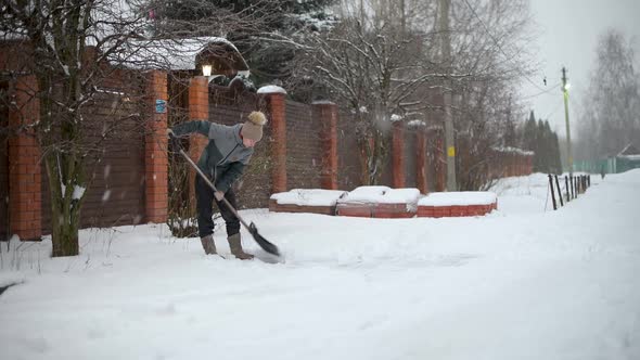 A young woman shovels snow in front of a private house on a winter evening