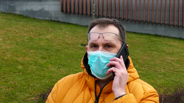 Sick Man in Medical Mask Speaks on the Phone While Sitting on a Park Bench