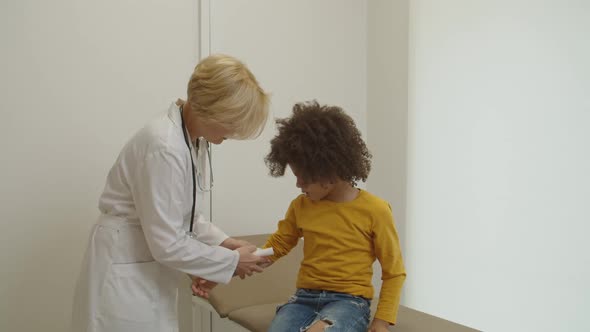 Female Physician Bandaging Injured Arm of Adorable Little Patient in Medical Clinic