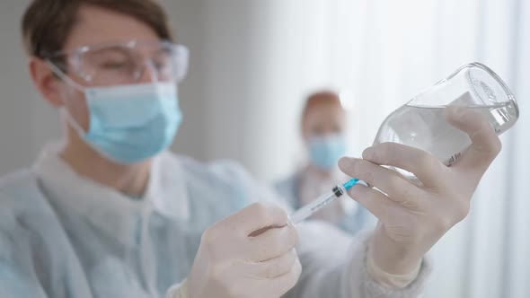 Male Doctor Filling Syringe with Liquid Medication As Blurred Nurse Standing at Background