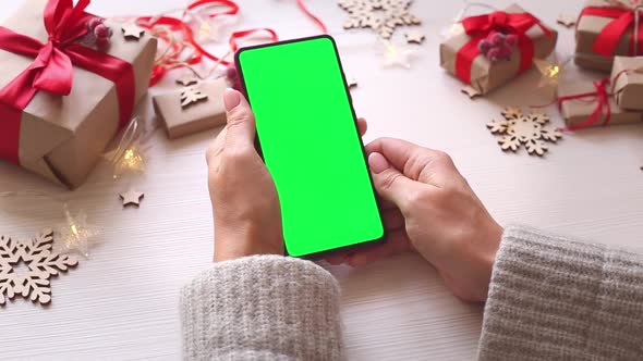 Woman holding smartphone with vertical green screen on Christmas background with gifts.