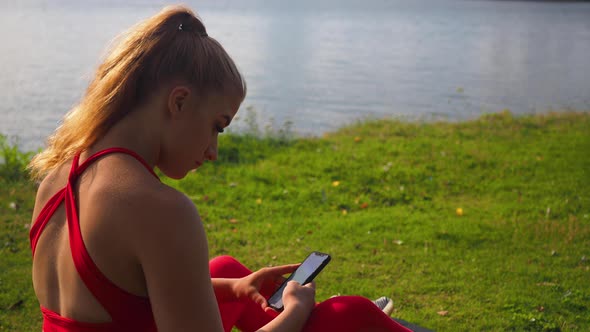 Young girl in red sportswear sitting on the grass at the seaside and scrolling on smartphone. View f