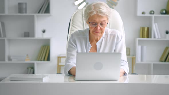 Senior lady secretary with grey hair and glasses in elegant suit types on laptop