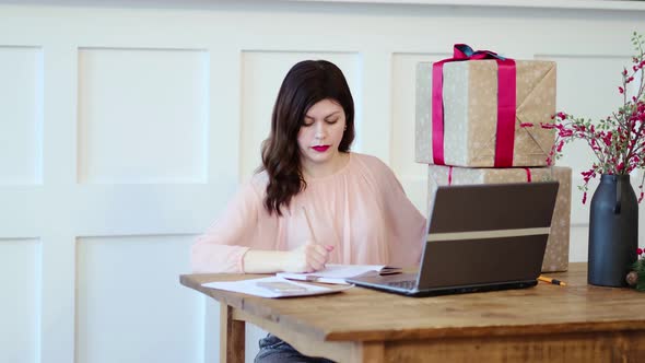 Pretty Young Lady Sitting at Desk and Typing on Laptop and Talking with Present Boxes Icons