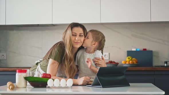 Little Girl Kisses and Whispers To Mom at Table in Kitchen