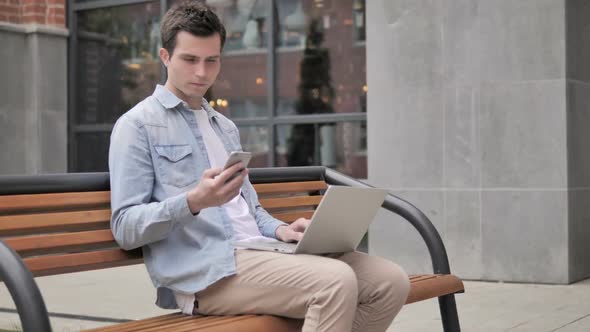 Young Man Sitting Outdoor Using Smartphone and Laptop