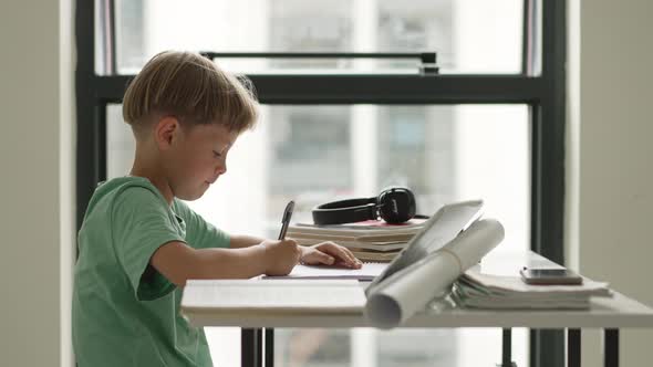 Caucasian Boy Sitting at Table with Workbook and Looking at Tablet Screen While Attentively