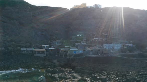 Aerial dolly over lava to village against the sun with rays in frame