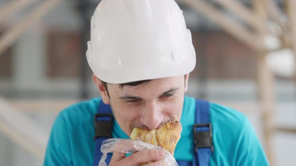 Portrait of Young Positive Service Man in Hard Hat Eating Sandwich Indoors