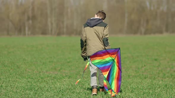 A Boy Walks Across the Field Carrying a Multicolored Kite in His Hands