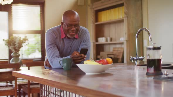 African american senior man leaning on kitchen counter using smartphone, taking off glasses, smiling