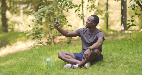 Cheerful African American Guy Talking on Mobile Phone, Sitting on Grass in Park