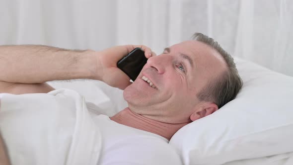 Positive Middle Aged Man Talking on Smartphone in Bed 