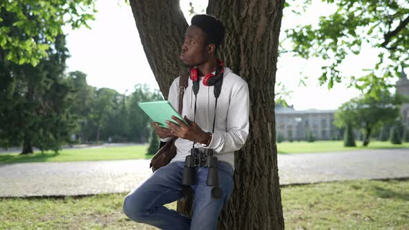 Young Focused African American Student Leaning on Tree Trunk Surfing Internet on Tablet