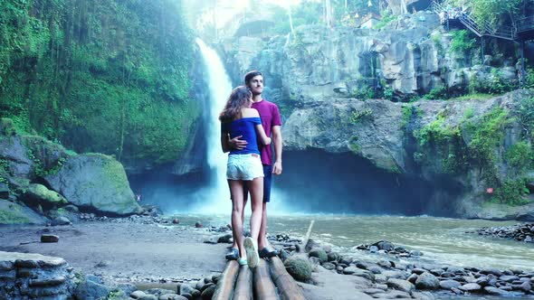 Hugging couple standing over bamboo pier, kissing in front of wonderful waterfall into valley with l