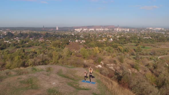 Aerial Orbiting Shot of Slender Young Mother Do Yoga Exercises with Child Daughter on High Hill By