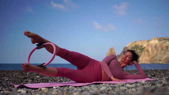 A Young Woman with Black Hair Doing Pilates with the Ring on the Yoga Mat Near the Sea on the Pebble