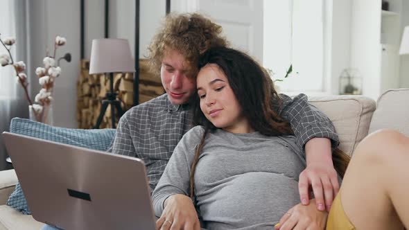 Husband and Pregnant Woman Relaxing Together on the Soft Couch in Living-Room and Using Computer