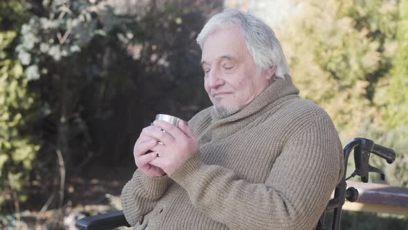 Side View Portrait of Elderly Caucasian Man Drinking Hot Tea As Sitting in Wheelchair Outdoors