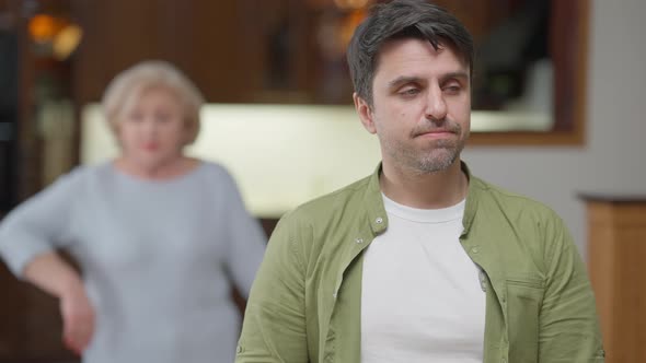 Portrait of Adult Caucasian Man Sighing As Angry Dissatisfied Senior Woman Arguing at Background