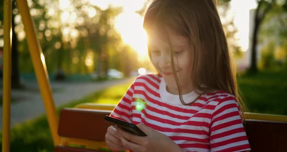 Portrait of Little Cheerful Blonde Girl Uses a Smartphone Outdoors