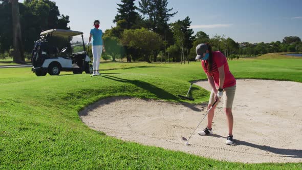 Two caucasian women playing golf wearing face masks one taking shot from bunker