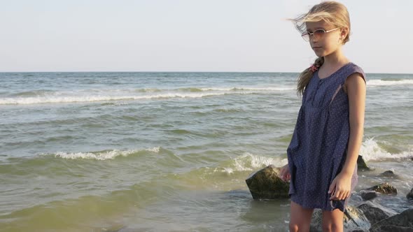 A Girl in a Blue Dress with Polka Dots and Sunglasses Looks Into the Distance on the Beach