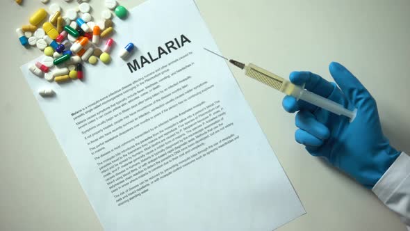Malaria Word Typed on Paper, Doctor Hand With Syringe Pills and Tablets on Table