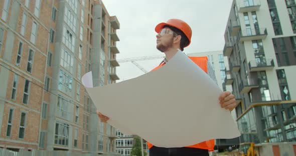 Young Architect or Builder With Hard Hat Holding Blueprint In His Hands in Front of Building Under