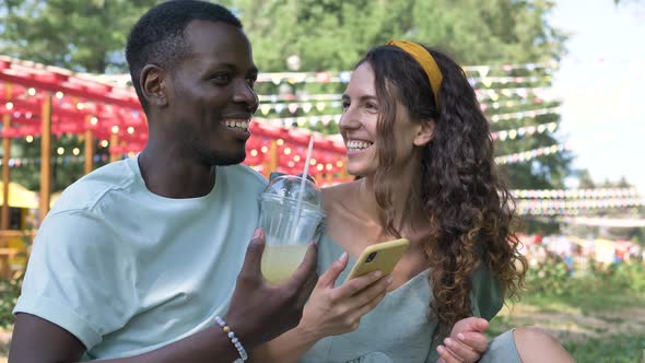 Happy Multiracial Couple in Love Drinking Cocktails in the Park and Chatting