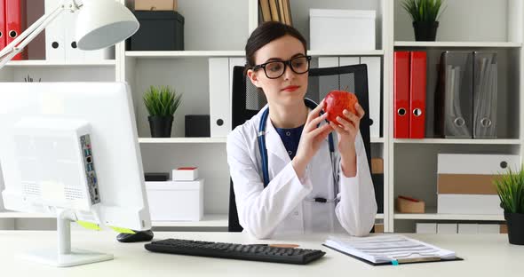 Doctor Sitting at Table, Looking at Red Apple and Making Gesture Okay