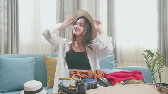 Asian Young Woman Try Hat And Packing Clothes In Suitcase At Home, Preparing For Vacation