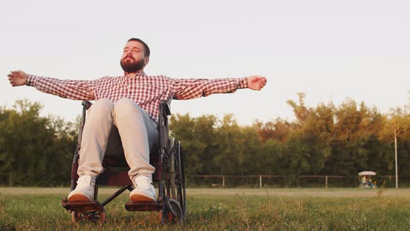 Disabled Hipster Sitting in Wheelchair on Field Against Background Sunset Spreads His Arms to Sides